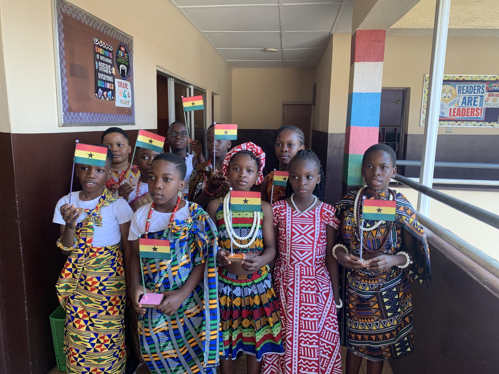 Pupils in kente and beads waving Ghanaian flags in a school corridor